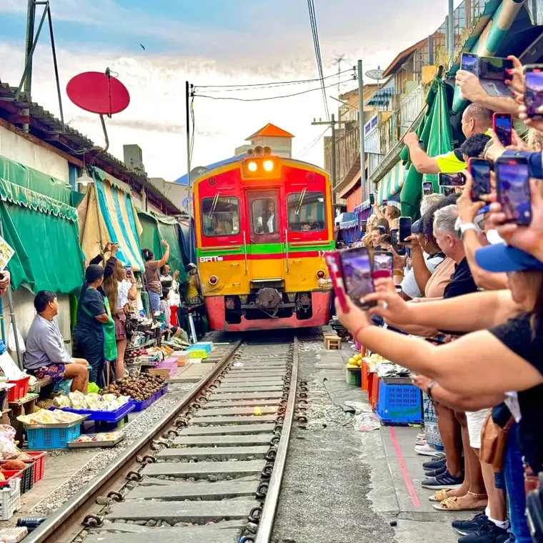 Train passing through the Maeklong Railway Market with visitors standing close to the tracks.